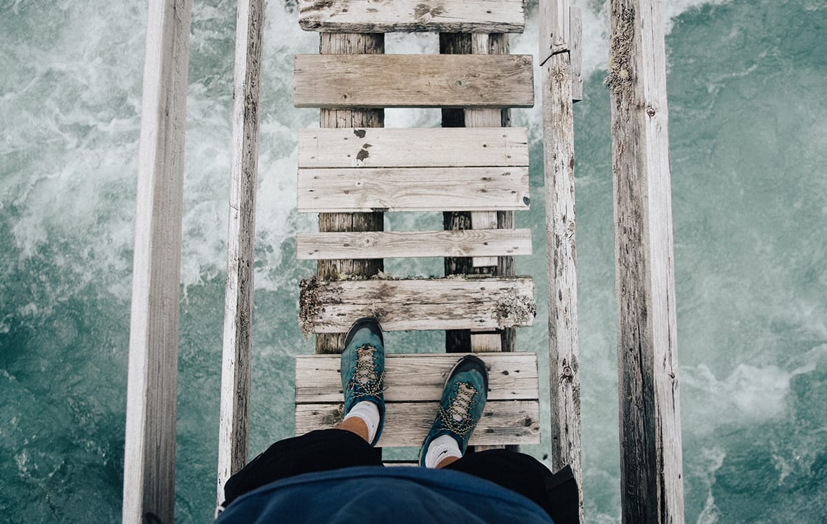 Person standing on a rickety wooden bridge looking down at the turbulent water below